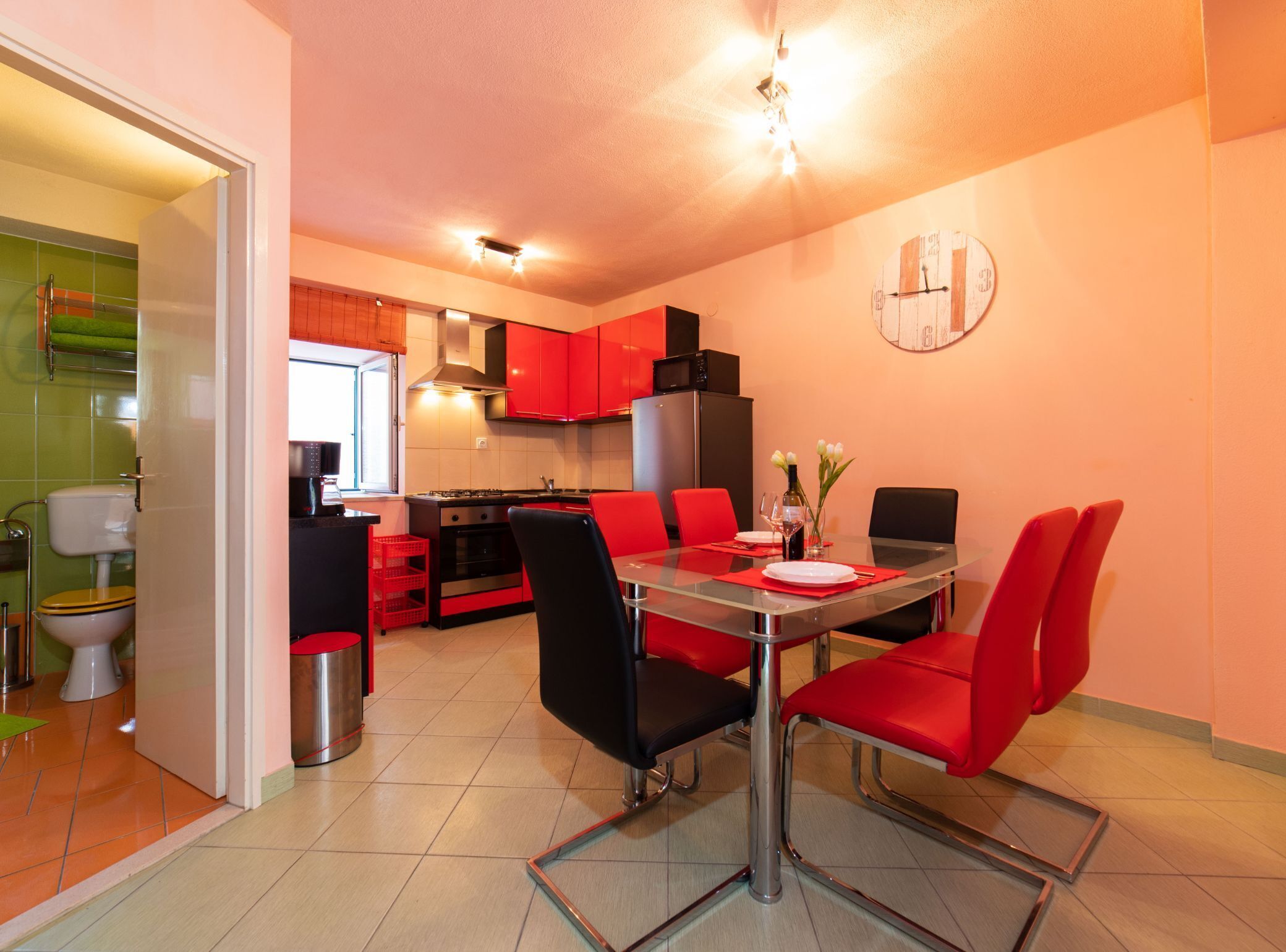 Una cocina compacta con armarios rojos, una mesa de comedor con sillas rojas y un reloj visible en la pared.