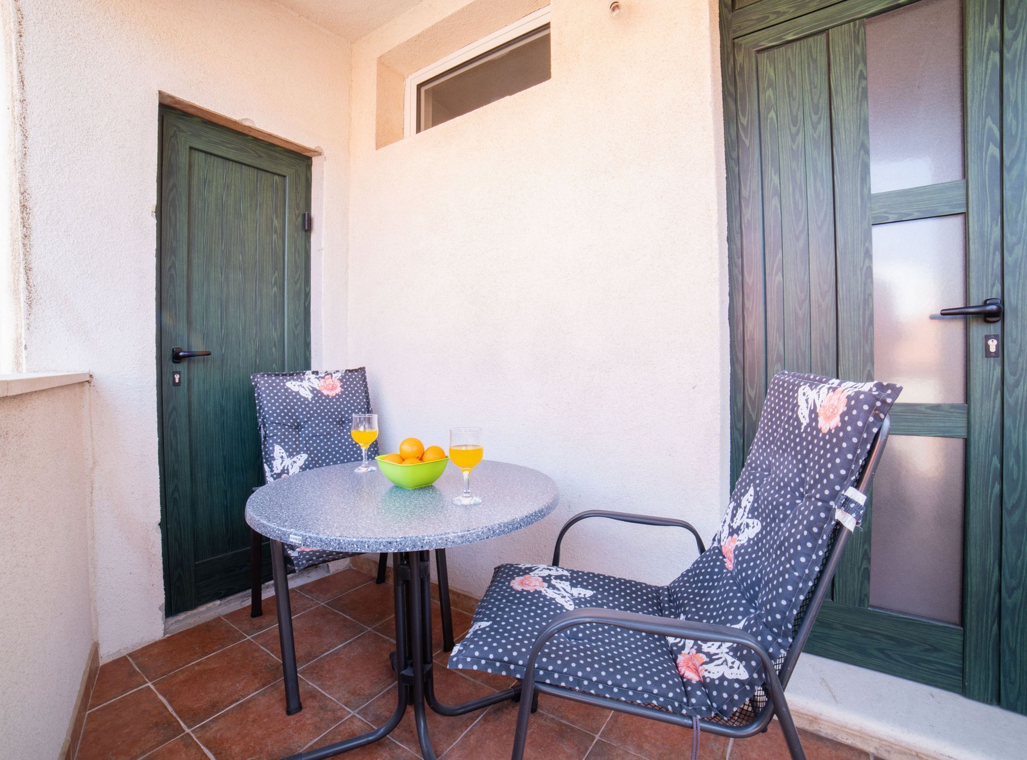 Un petit espace patio avec une table ronde et deux chaises, un saladier rempli de fruits et deux verres de jus d’orange.