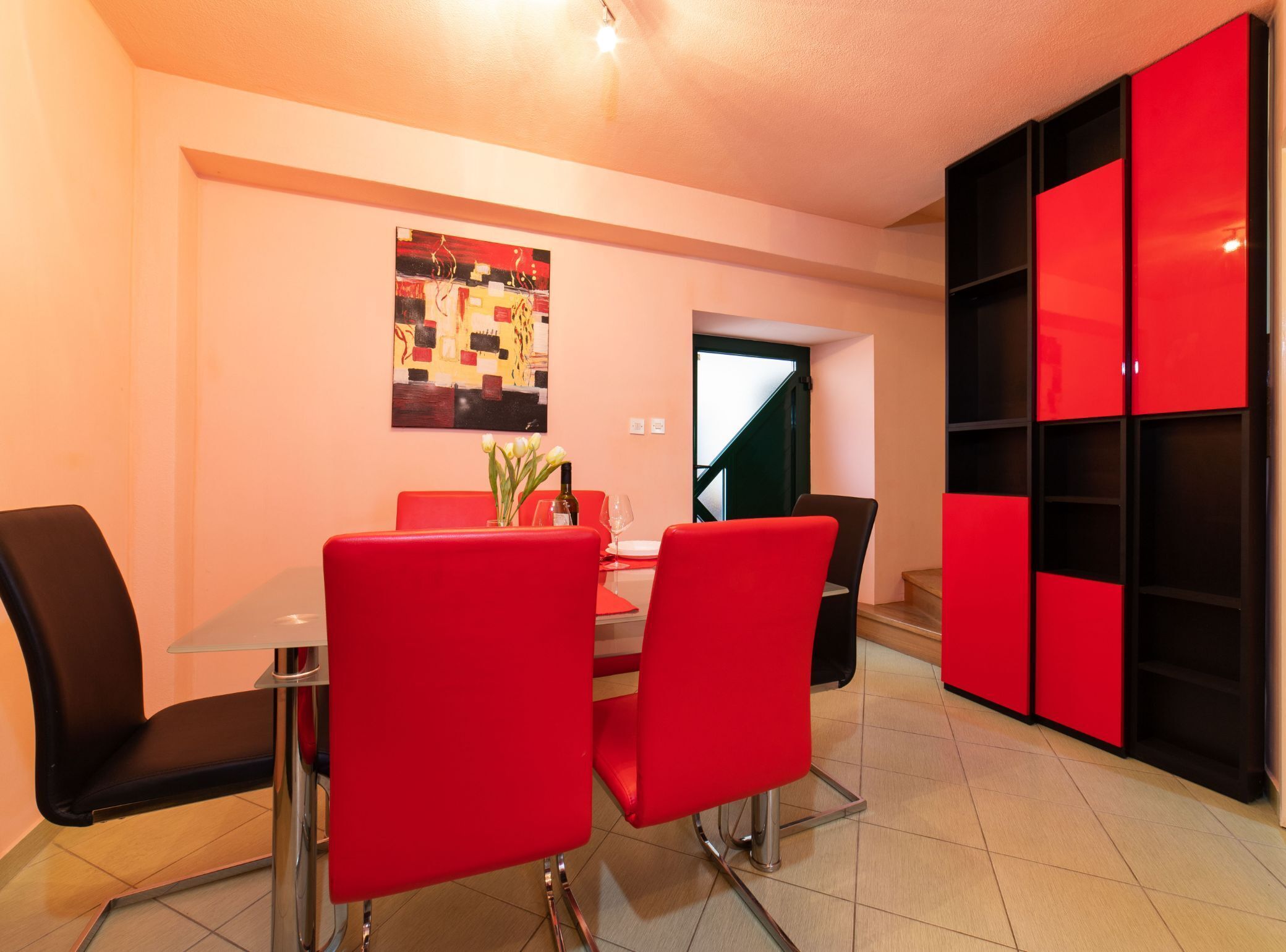 Un comedor con mesa de vidrio, sillas rojas, un cuadro en la pared y una escalera que lleva a otra habitación.