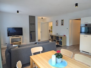 Un salon moderne avec un téléviseur à écran plat, un canapé gris, une table à manger en bois et une petite plante en pot.