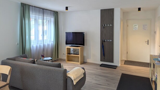 Ein modernes Wohnzimmer mit einer Couch, einem Fernseher auf einem Schrank, einem kleinen Tisch mit einer Vase und einer Tür mit einem Schild.