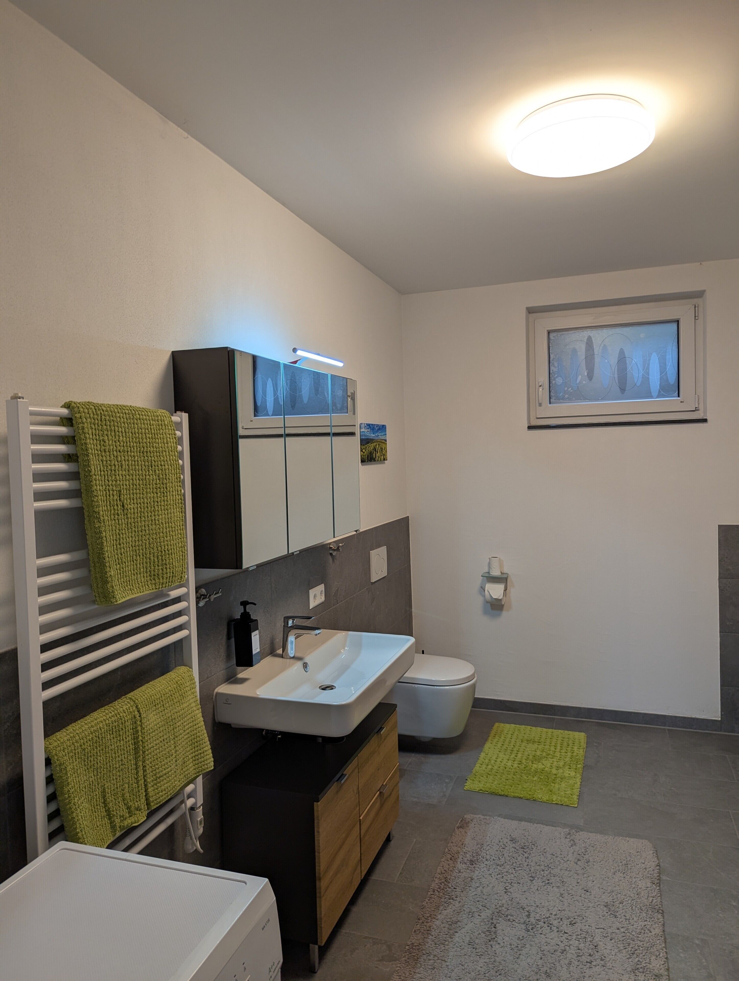 A modern bathroom with a white sink, a mirror, a towel rack, a toilet, and a small window with a painting.