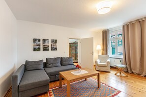 Una sala de estar con un sofá gris, una mesa de centro de madera, un tapiz estampado, un sillón blanco y una ventana con cortinas.