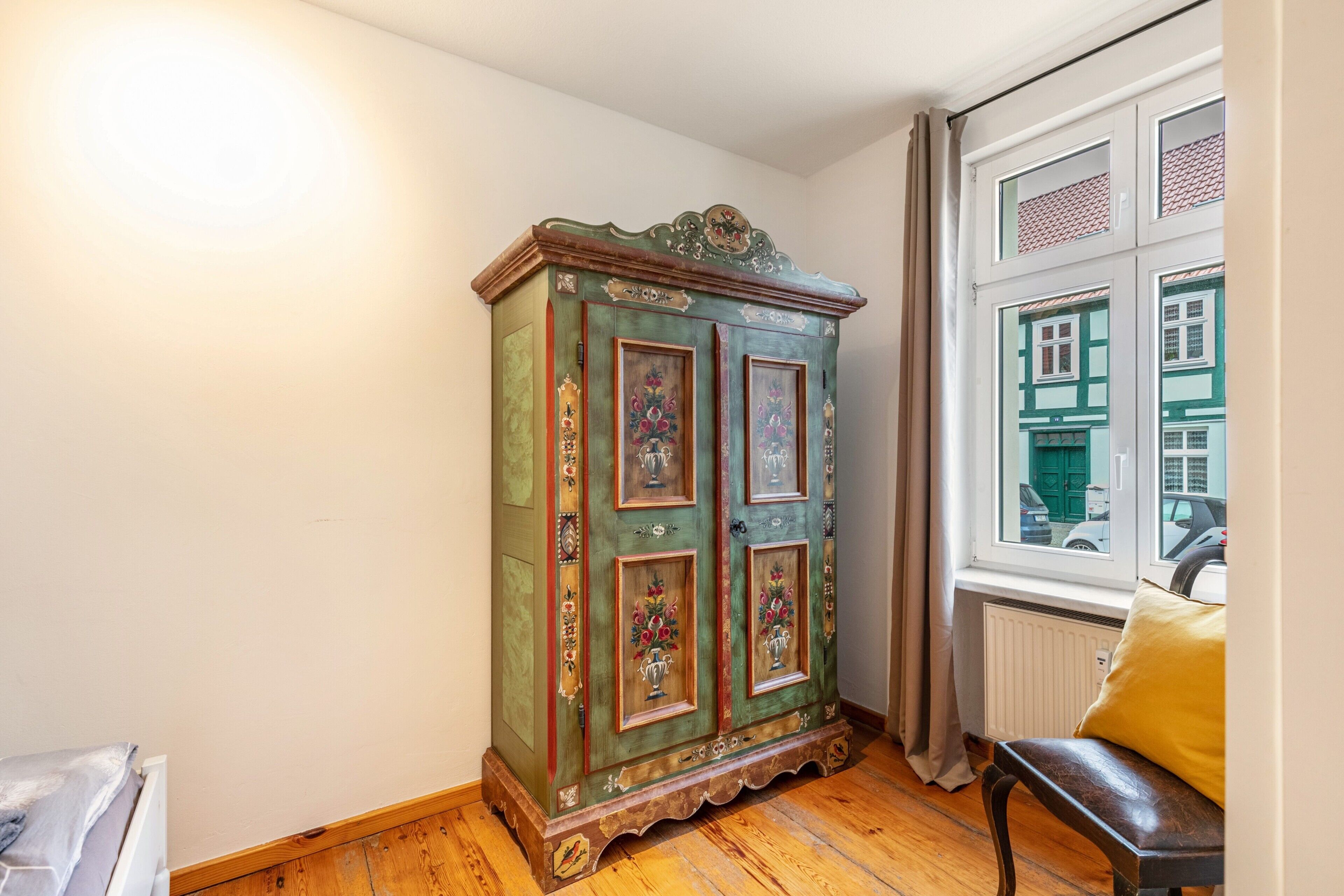 Une pièce avec un plancher en bois, un meuble vert orné de motifs floraux, une fenêtre donnant sur des immeubles et un coussin jaune posé sur un banc.