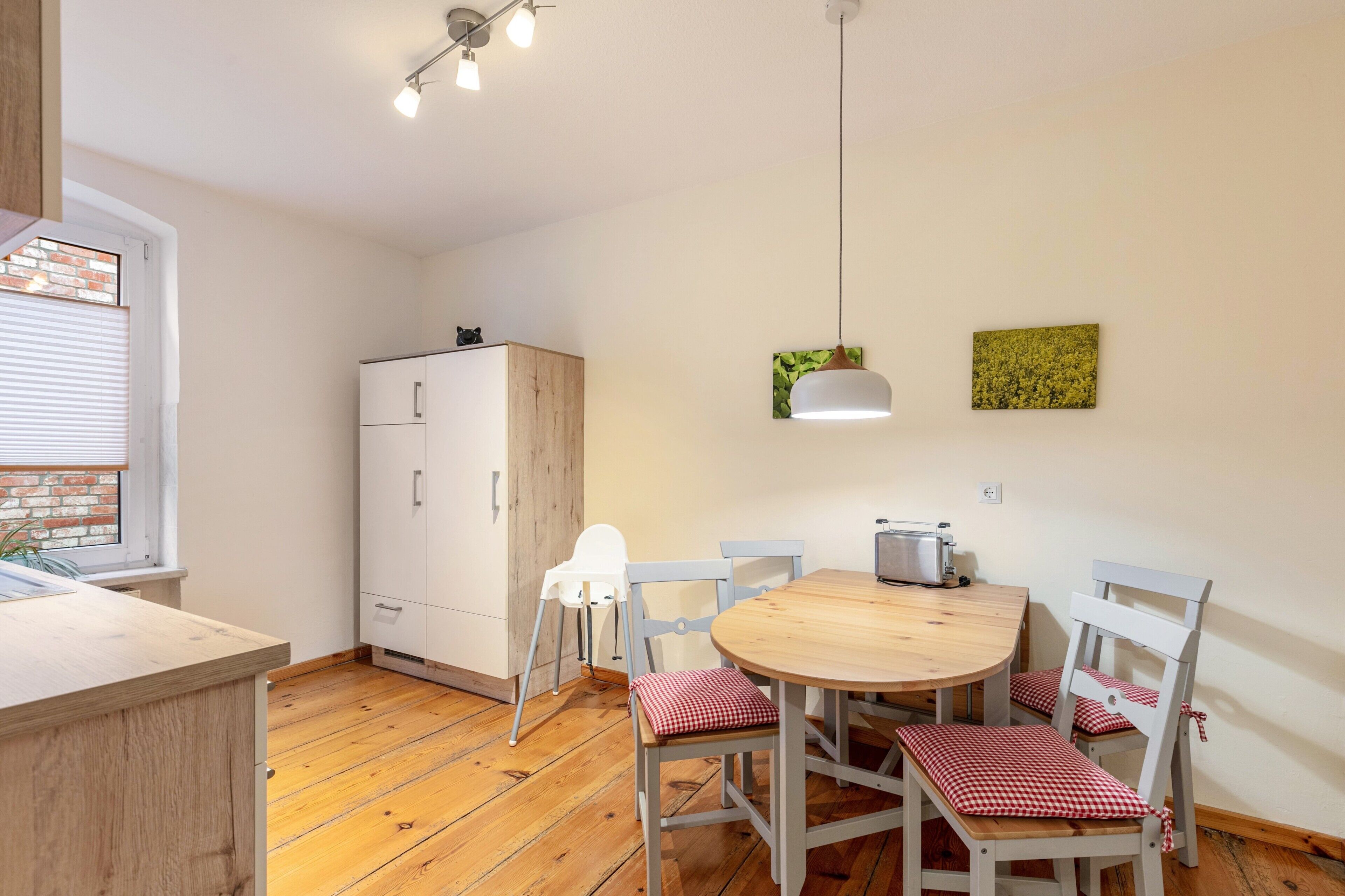 A compact kitchen with a wooden countertop, a white cabinet, a round wooden table with four chairs, a toaster, and a hanging light fixture.