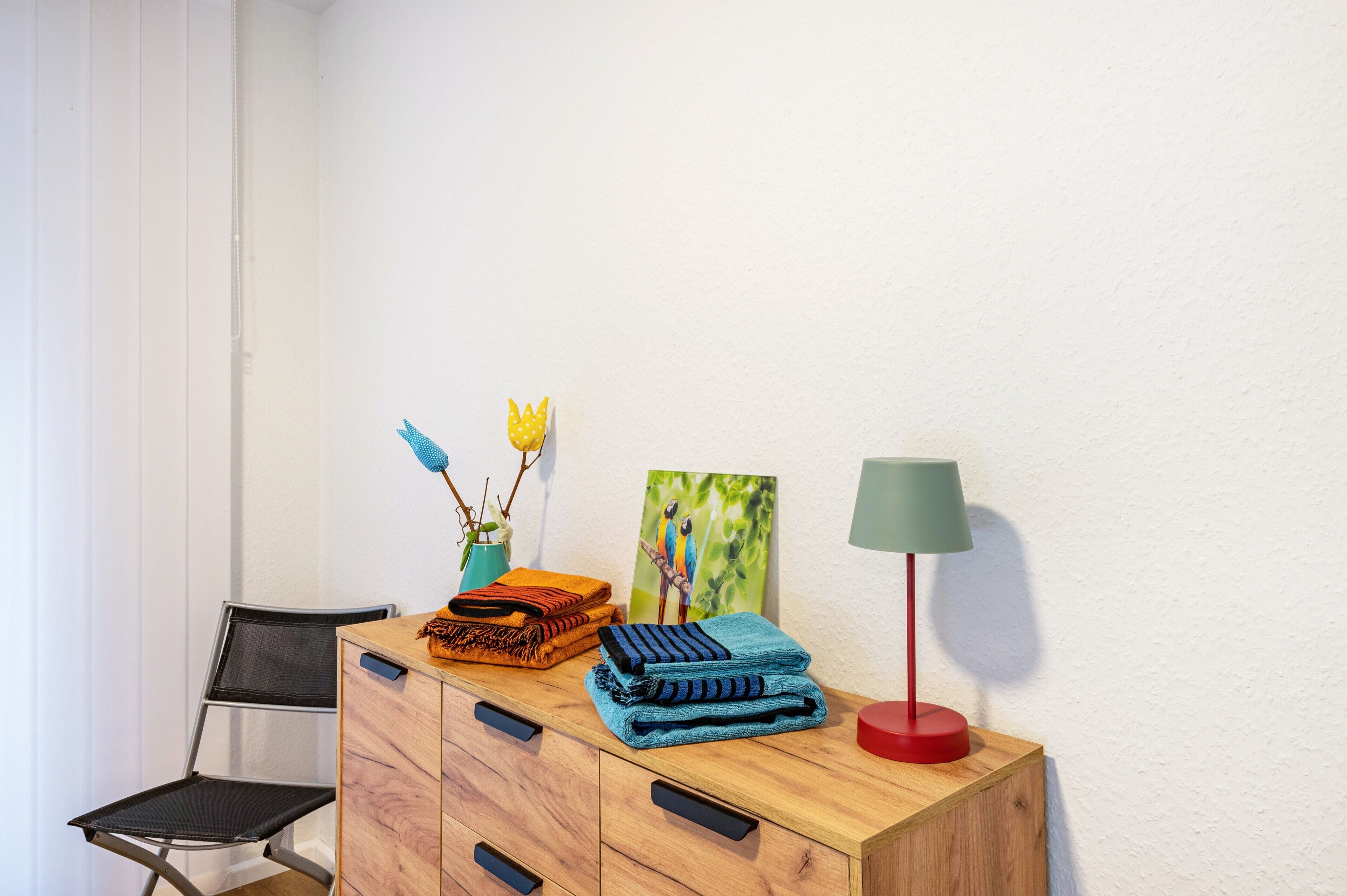 Une commode en bois, avec des couvertures pliées, une lampe, un tableau et une chaise.