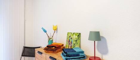 Une commode en bois, avec des couvertures pliées, une lampe, un tableau et une chaise.
