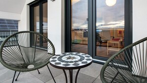 A balcony with two wicker chairs and a small tiled table, large glass doors, and a view of the cityscape.