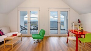 A modern loft apartment with a high ceiling, a dining area with a red table and green chairs, a green armchair, a white sofa, and a balcony with a view.