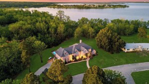 A large house with a stone exterior, surrounded by a well-maintained lawn and trees, near a lake. - The Cove: Heated Pool, Hot Tub, Lake Access (Shady Shores)