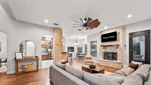 A modern living room with a stone fireplace, a large gray sofa, and a dining area in the background. - The Cove: Heated Pool, Hot Tub, Lake Access (Shady Shores)