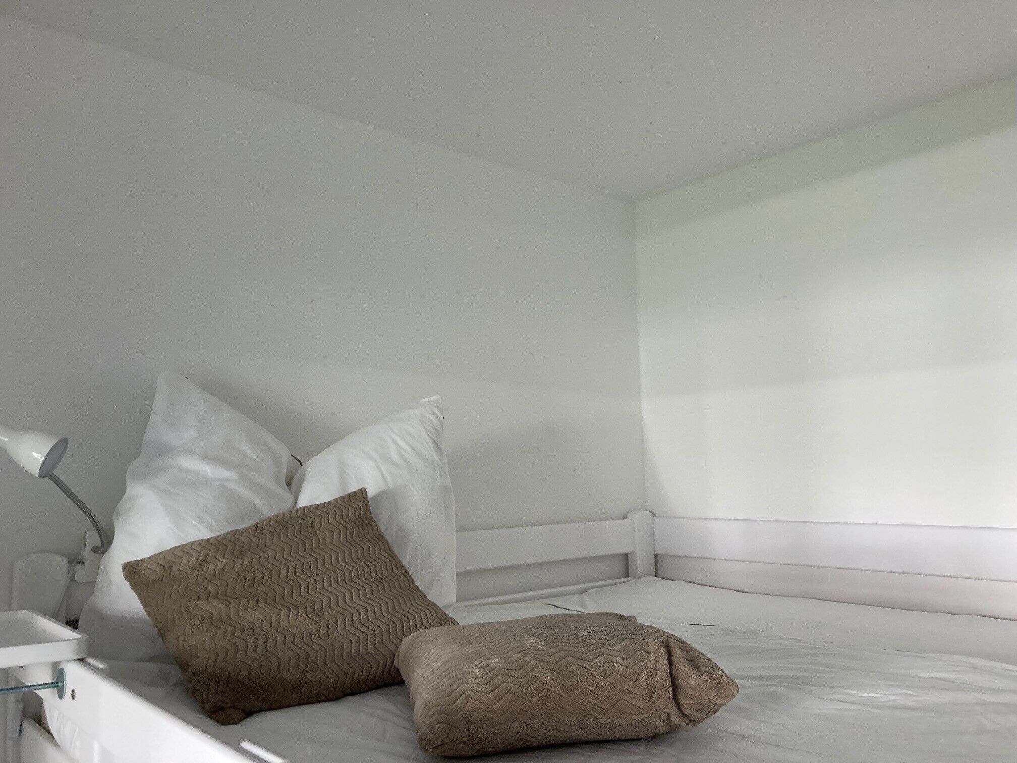 Ein weißes Bett mit zwei Kissen, ein Nachttisch mit Lampe und ein Fenster mit Jalousien.