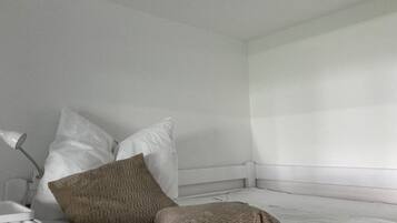 Ein weißes Bett mit zwei Kissen, ein Nachttisch mit Lampe und ein Fenster mit Jalousien.