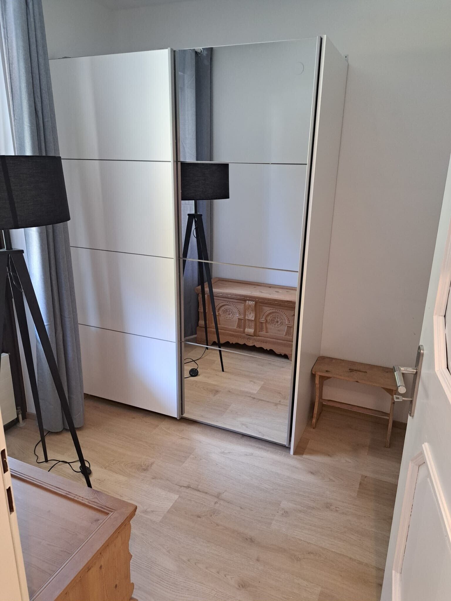 Ein Zimmer mit Schiebetürenschrank, einer kleinen Holzbank und einer auf einem Stativ stehenden Lampe.