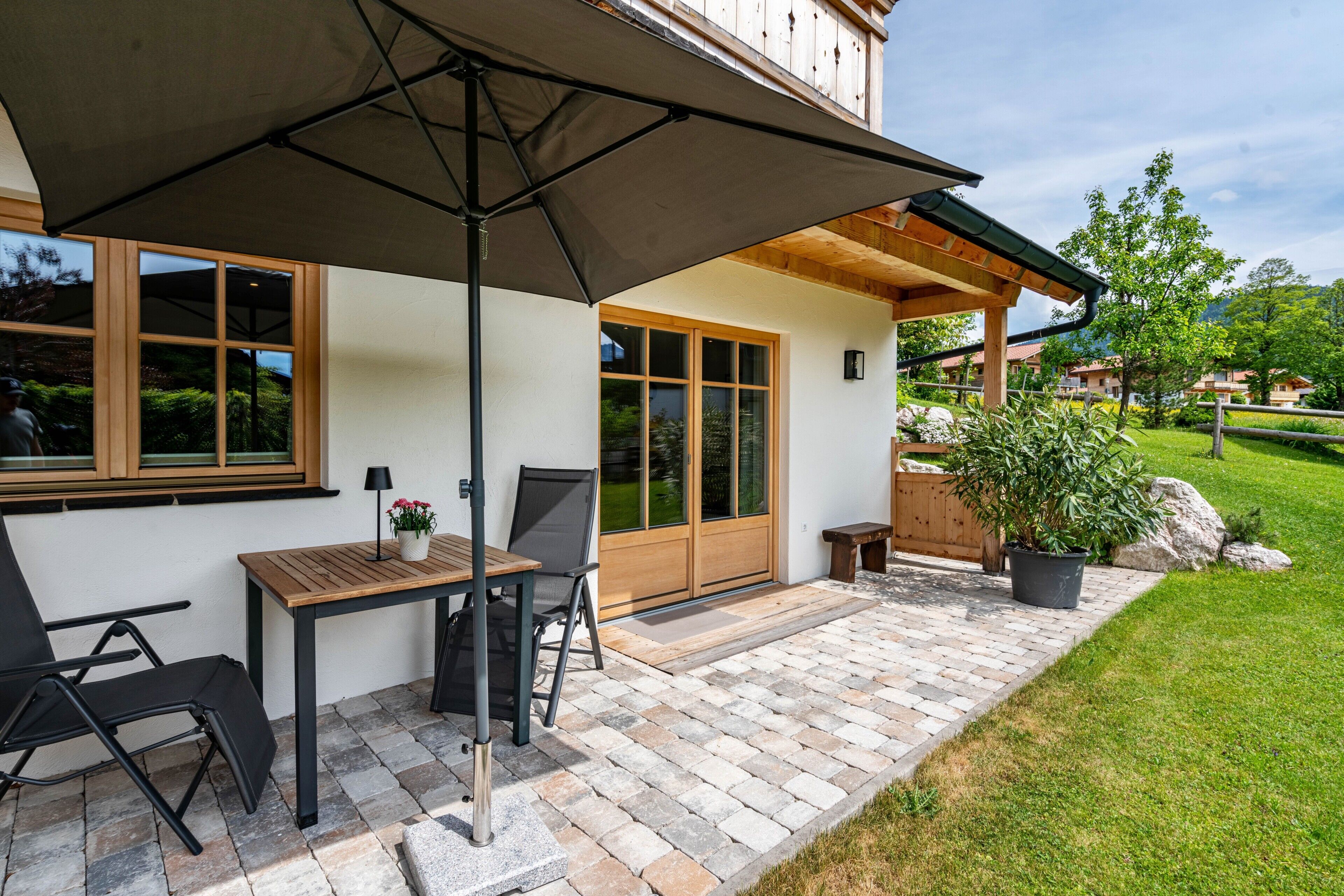 Eine Terrasse mit Tisch und Stühlen, ein Holzboden und Blick auf ein Gebäude mit großen Fenstern.