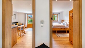 Overzicht van een kamer met een houten vloer, een bed met kussens, een bureau met stoel en een raam met uitzicht op groen.