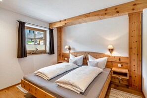 Cama doble con cabecera de madera, dos mesitas de noche con lámparas, una ventana con cortinas y vistas a un edificio.