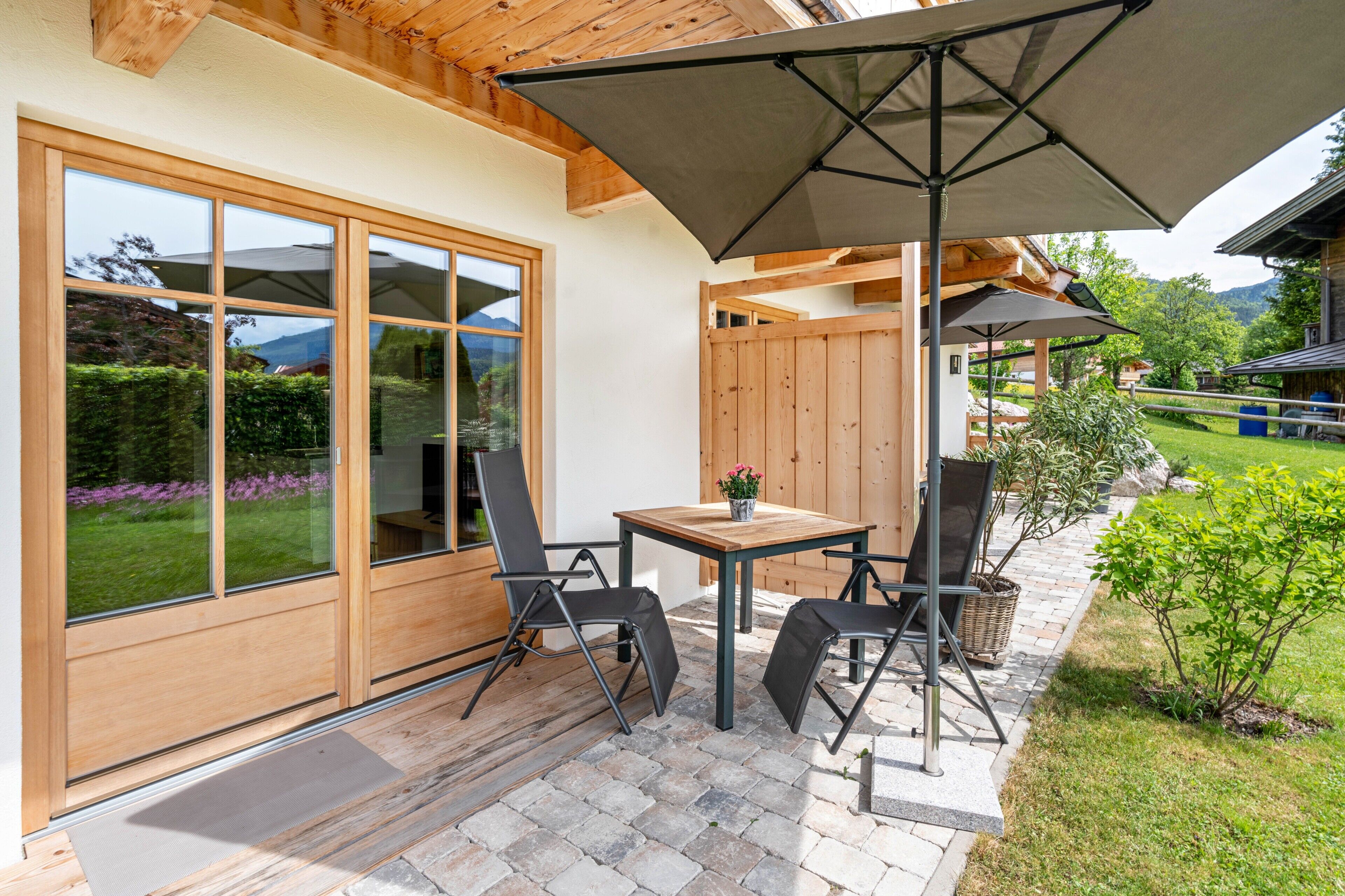 A patio with a table and chairs, a wooden deck, and a large window with a view of greenery and mountains.