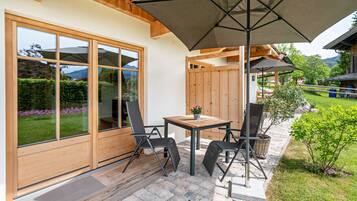 A patio with a table and chairs, a wooden deck, and a large window with a view of greenery and mountains.