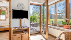 A room with a television, wooden furniture, a window with a view of greenery, and a door leading outside.