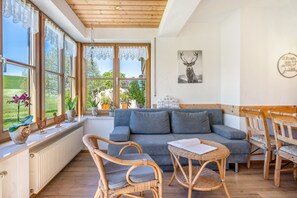 Une pièce chaleureuse avec un canapé gris, des fauteuils en osier, une petite table et une vue sur la verdure à l’extérieur.