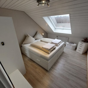 Une chambre avec un plafond en pente, un puits de lumière, un lit doté de tiroirs de rangement, un radiateur et une petite armoire blanche.