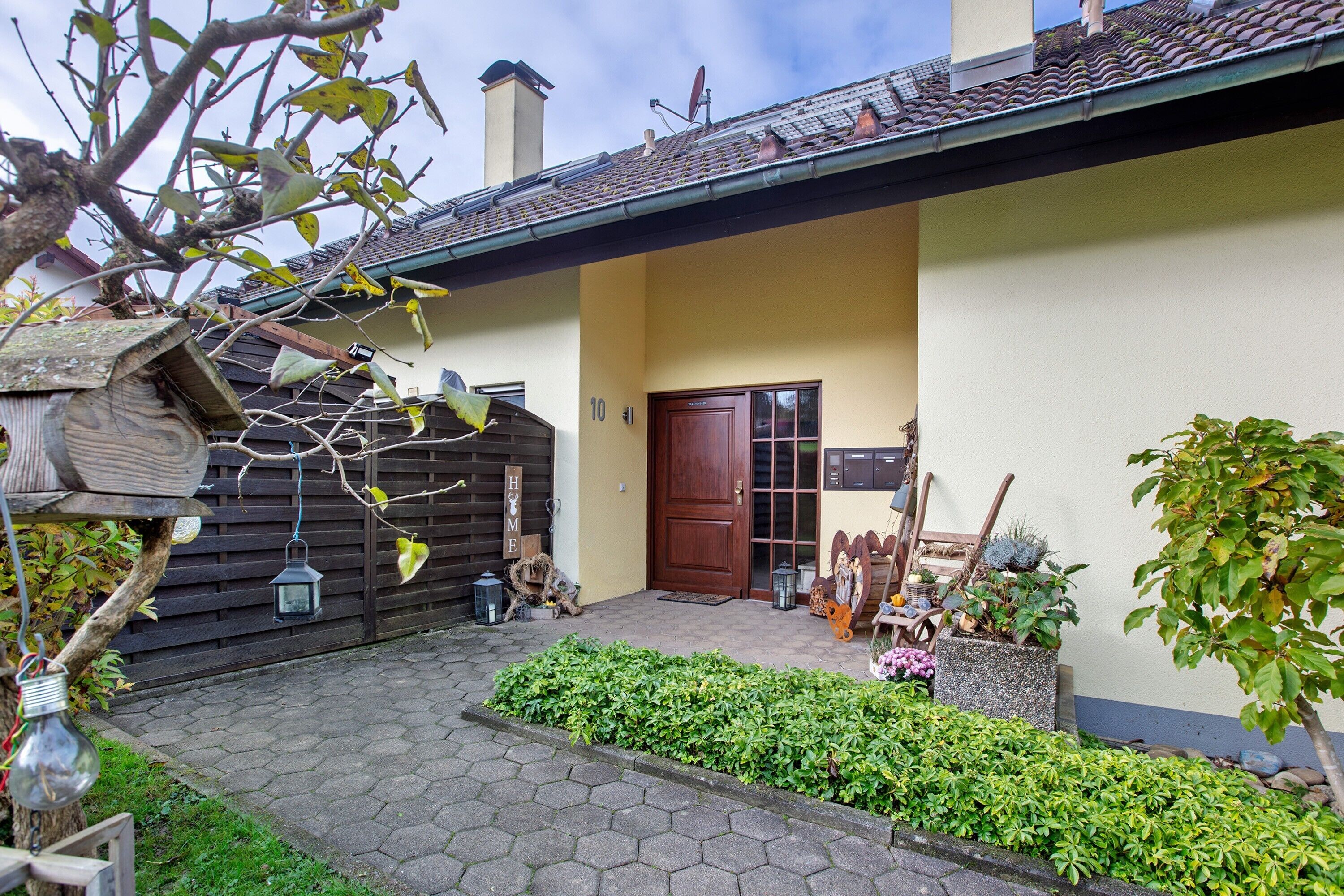 Une maison avec un toit en tuiles, une porte en bois et un jardin agrémenté de plantes et d’une nichoir.