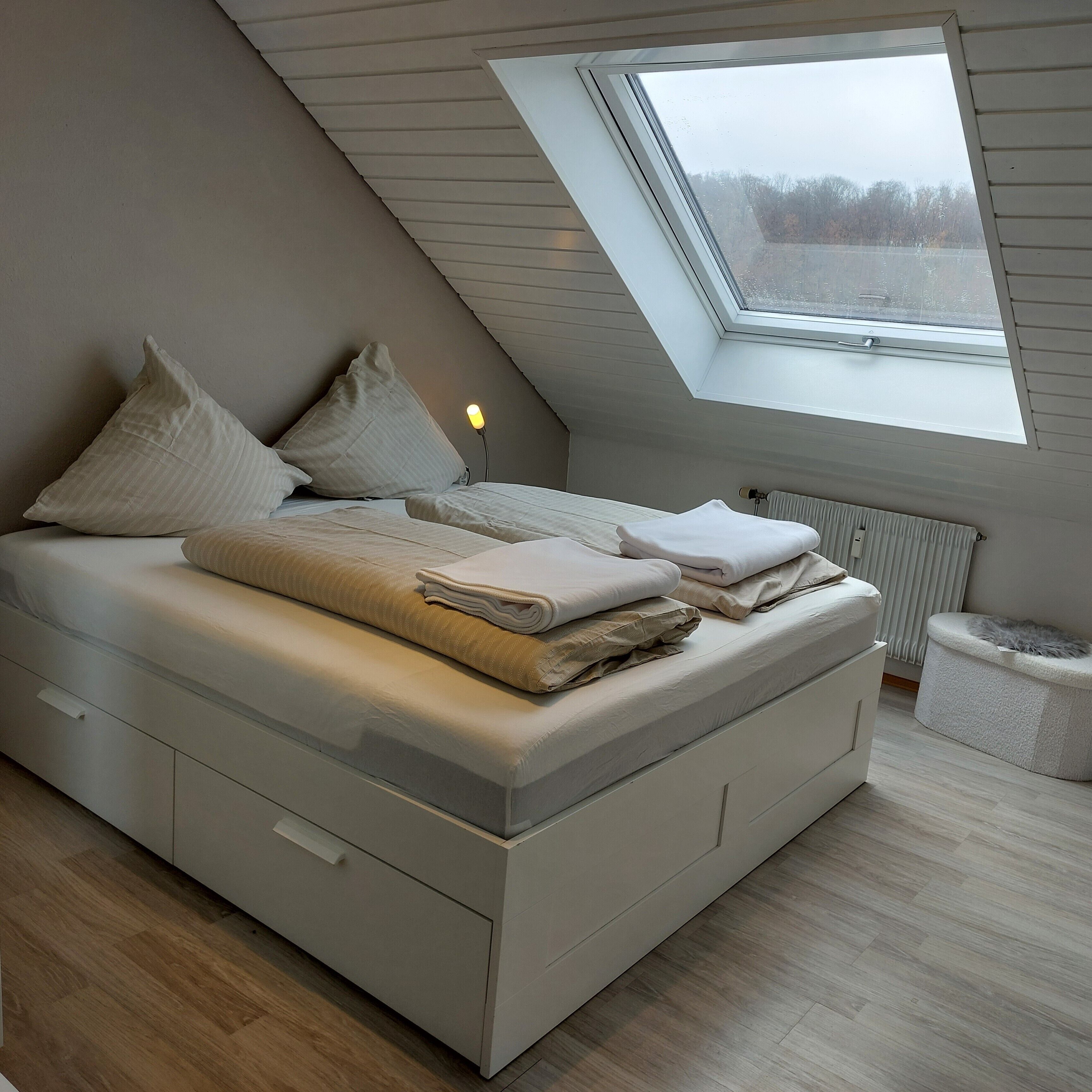 Ein modernes Schlafzimmer im Loft-Stil mit schrägen Wänden, einem Oberlicht, einem Bett mit Kissen und Decken, einem Nachttisch mit Lampe und einer Heizung.
