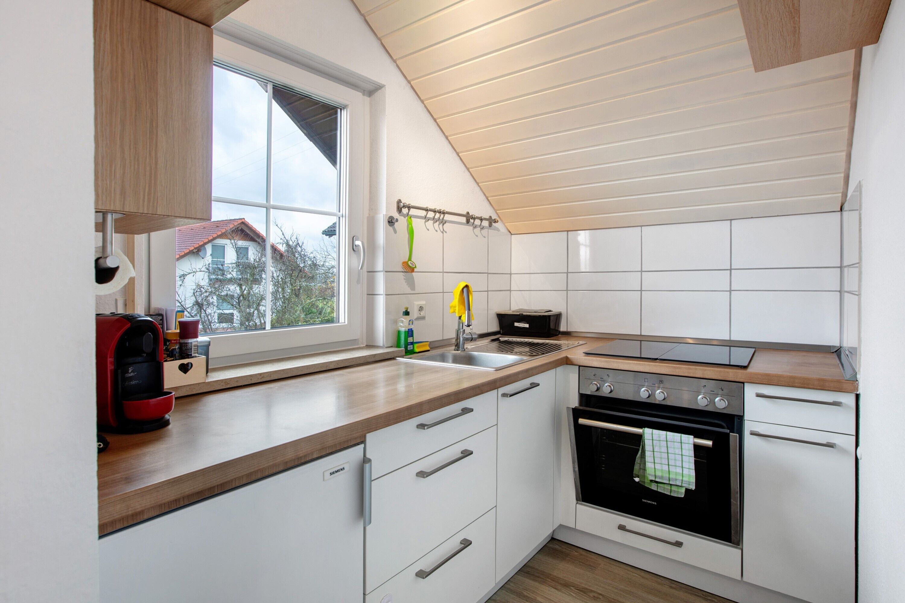 Une cuisine moderne avec des meubles blancs, un four encastré, une machine à café et une fenêtre donnant sur une maison.