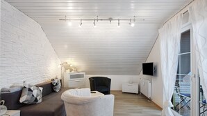A modern loft bedroom with a sloped ceiling, a white brick wall, a large window with curtains, a flat-screen TV, and a cozy seating area with a sofa and armchair. - Holiday Apartment 'Lueg Ins Wiesental' with Mountain View, Balcony & Wi-Fi (Steinen)