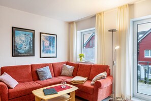 Un sofá rojo, una mesita de madera y una lámpara de pie en una habitación con una ventana y dos cuadros en la pared.