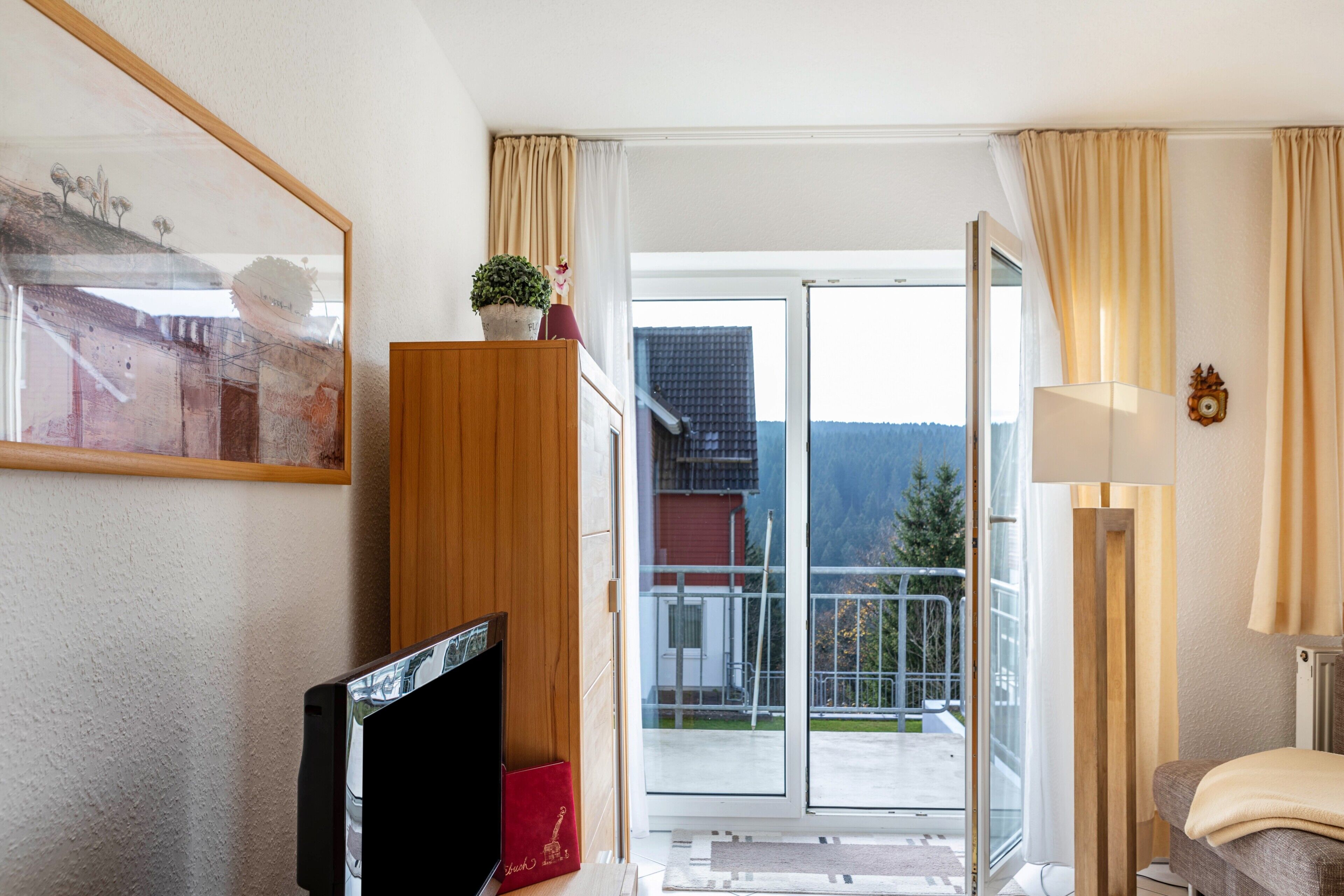 A hotel room with a television, a wooden cabinet, a balcony with a view of the mountains, and a lamp.