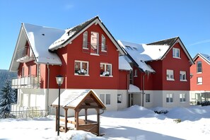 A red multi-story building with balconies, a wooden structure in the foreground, and snow on the roof.