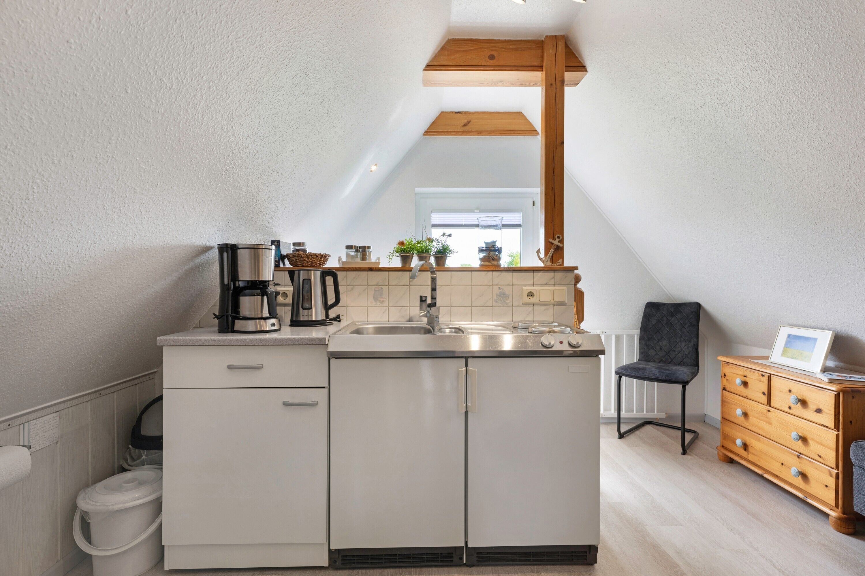 Una cocina compacta con estufa integrada, pileta y muebles blancos. Se observa una silla de madera y un pequeño aparador de madera con cajones.