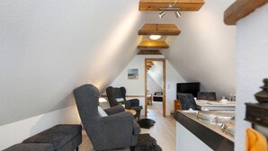 Un espacio tipo loft con techo alto, vigas de madera, un sofá, una silla, una pequeña mesa y vistas a través de un vano abierto.