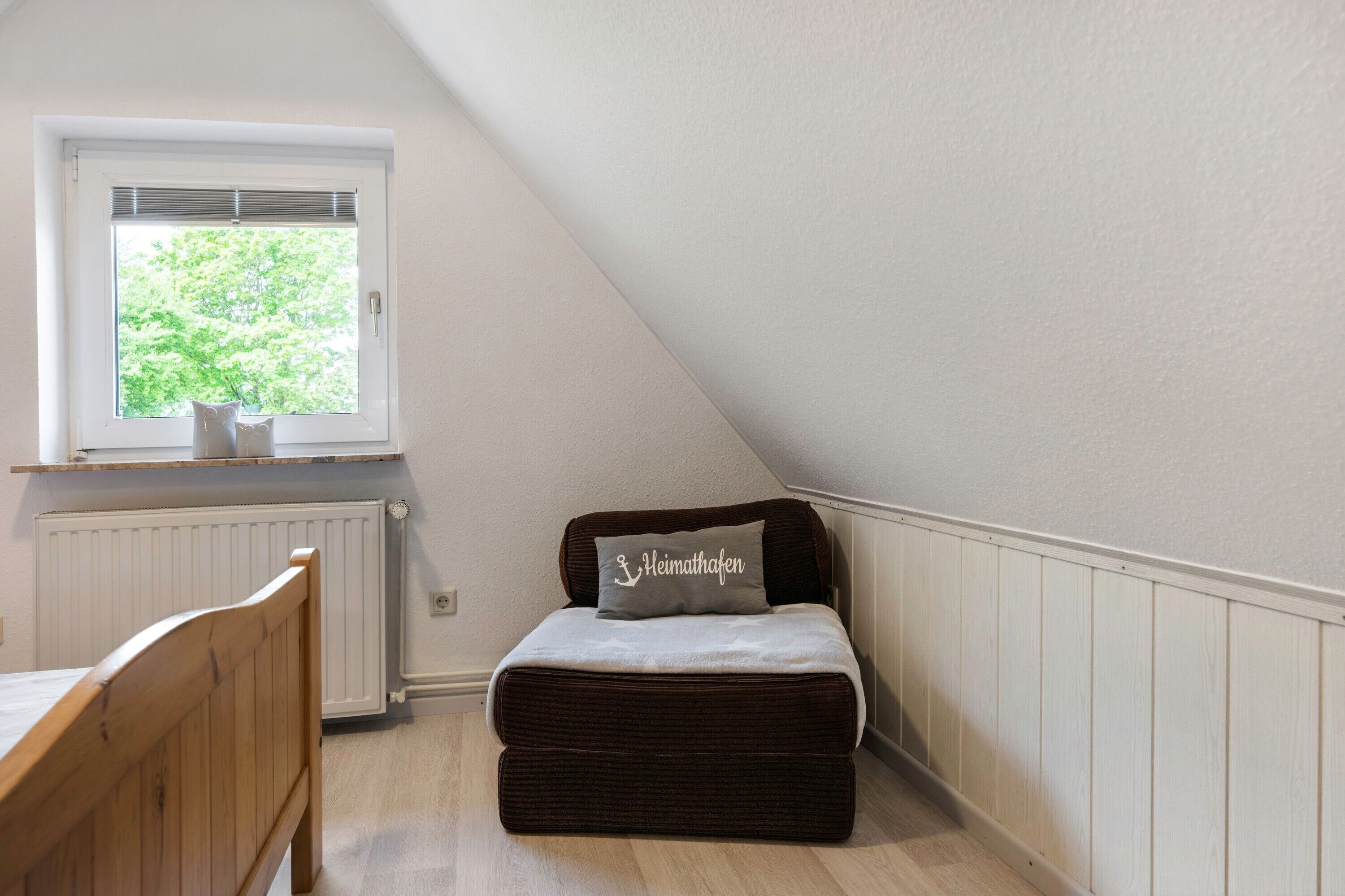 Une petite pièce agréable, dotée d’un plafond en pente, d’une fenêtre avec des stores, d’un radiateur, d’un lit en bois et d’un banc brun foncé agrémenté d’un coussin.