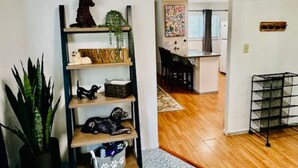 Un salon avec une bibliothèque, une plante en pot et un coin cuisine visible à travers une porte ouverte.