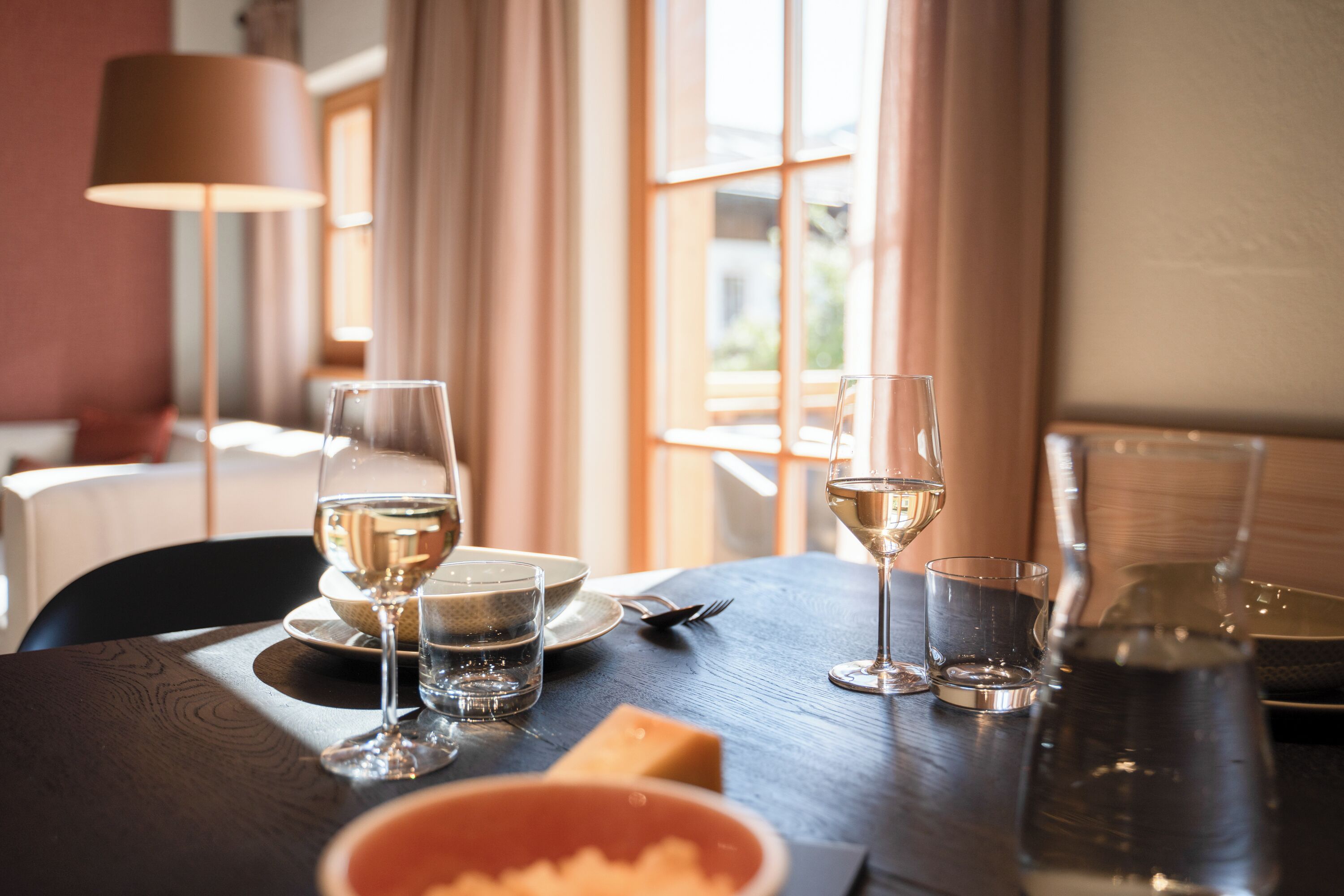 Een eettafel gedekt met wijnglazen, borden en een schaal eten, met een lamp en gordijnen op de achtergrond.