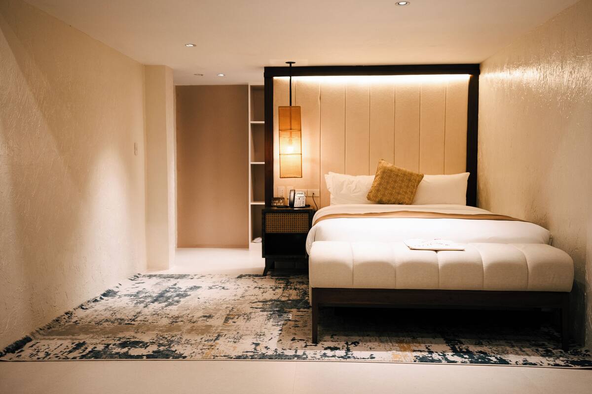 A modern hotel room with a large bed, a bedside table, a nightstand, and a wall-mounted lamp.
