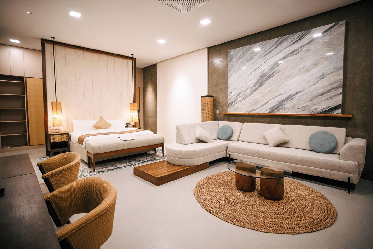 A modern hotel room with a bed, sofa, wicker chairs, a glass coffee table, and a large marble wall.