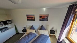 A bedroom with a bed, two bedside tables, a nightstand, a radiator, a window with curtains, and two wall-mounted pictures.