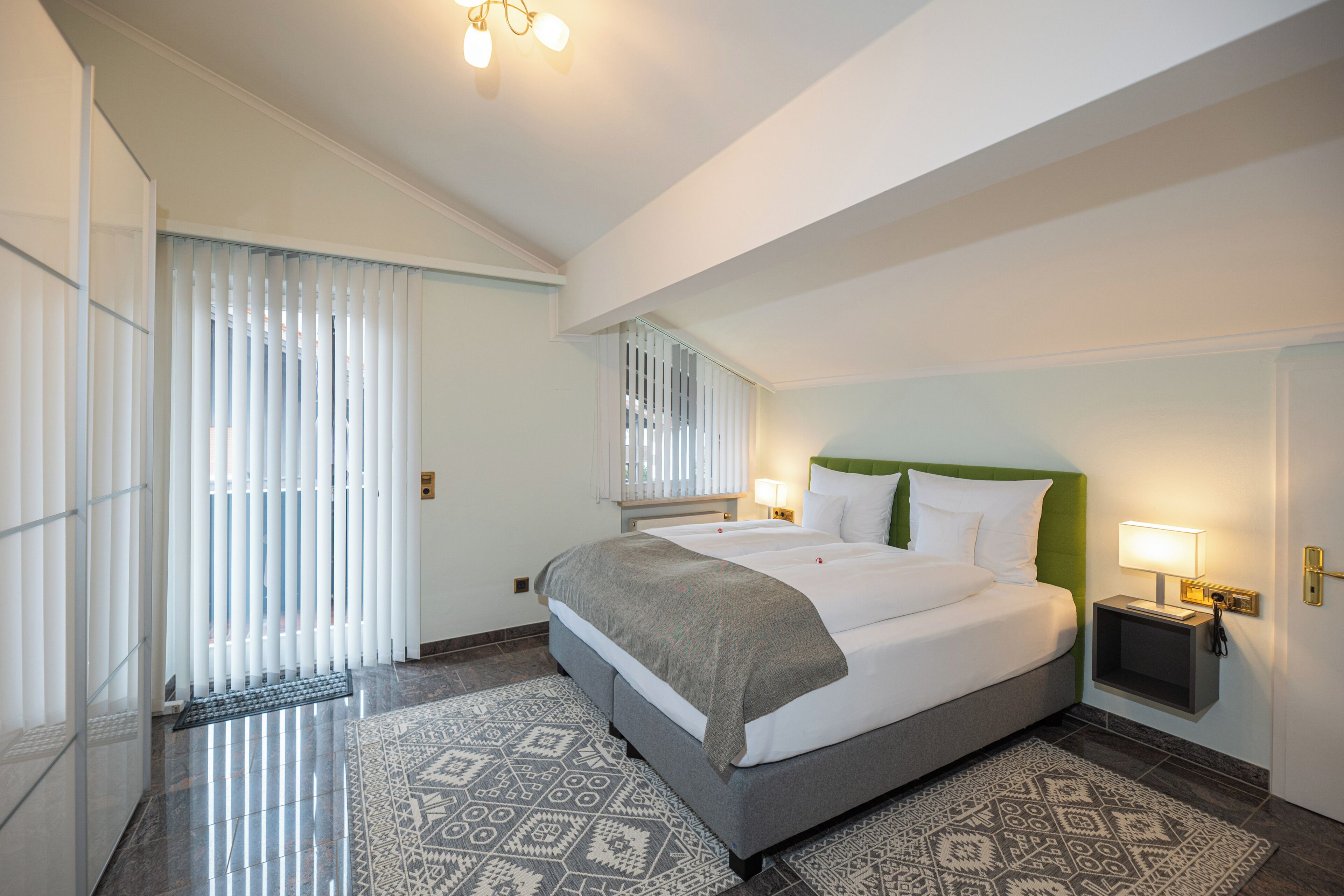 Una habitación de hotel con una cama grande, una mesita de noche con lámpara, un tapete estampado y una ventana con persianas.