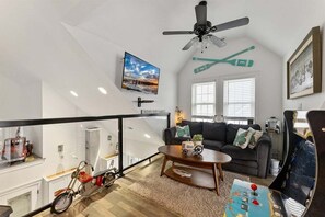 Una sala de estar con ventilador de techo, televisor montado, sofá con almohadones decorativos, una mesa de centro y una bicicleta en la esquina.