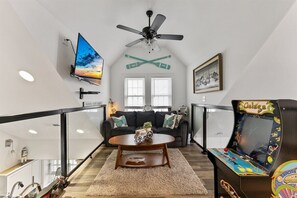 A living room with a flat-screen TV, ceiling fan, couch, coffee table, and arcade game.