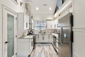 Una cocina con muebles blancos, electrodomésticos de acero inoxidable y una ventana con persianas.