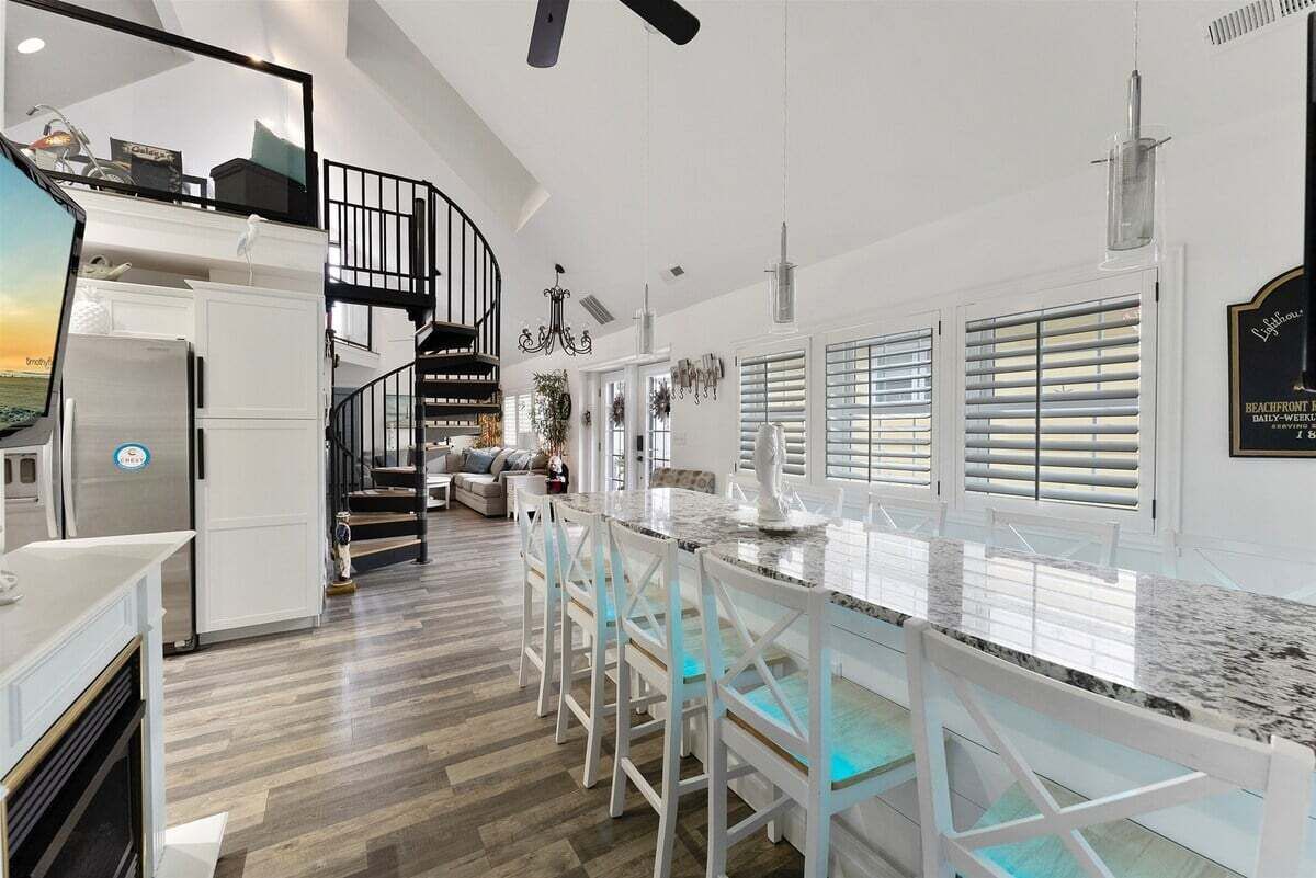 Una cocina moderna con una isla central, taburetes blancos en la barra, una escalera y vistas a la playa.
