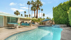 A swimming pool with lounge chairs and umbrellas, surrounded by palm trees and a building with a light-colored exterior. - Azul Oasis by AvantStay | Pool, Hot Tub, Firepit, Bocce Ball | Mins to Downtown (Palm Springs)