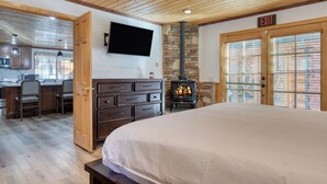 Una habitación amplia con una cama grande, techo de madera, chimenea de piedra, televisor y una zona de cocina con taburetes de bar.