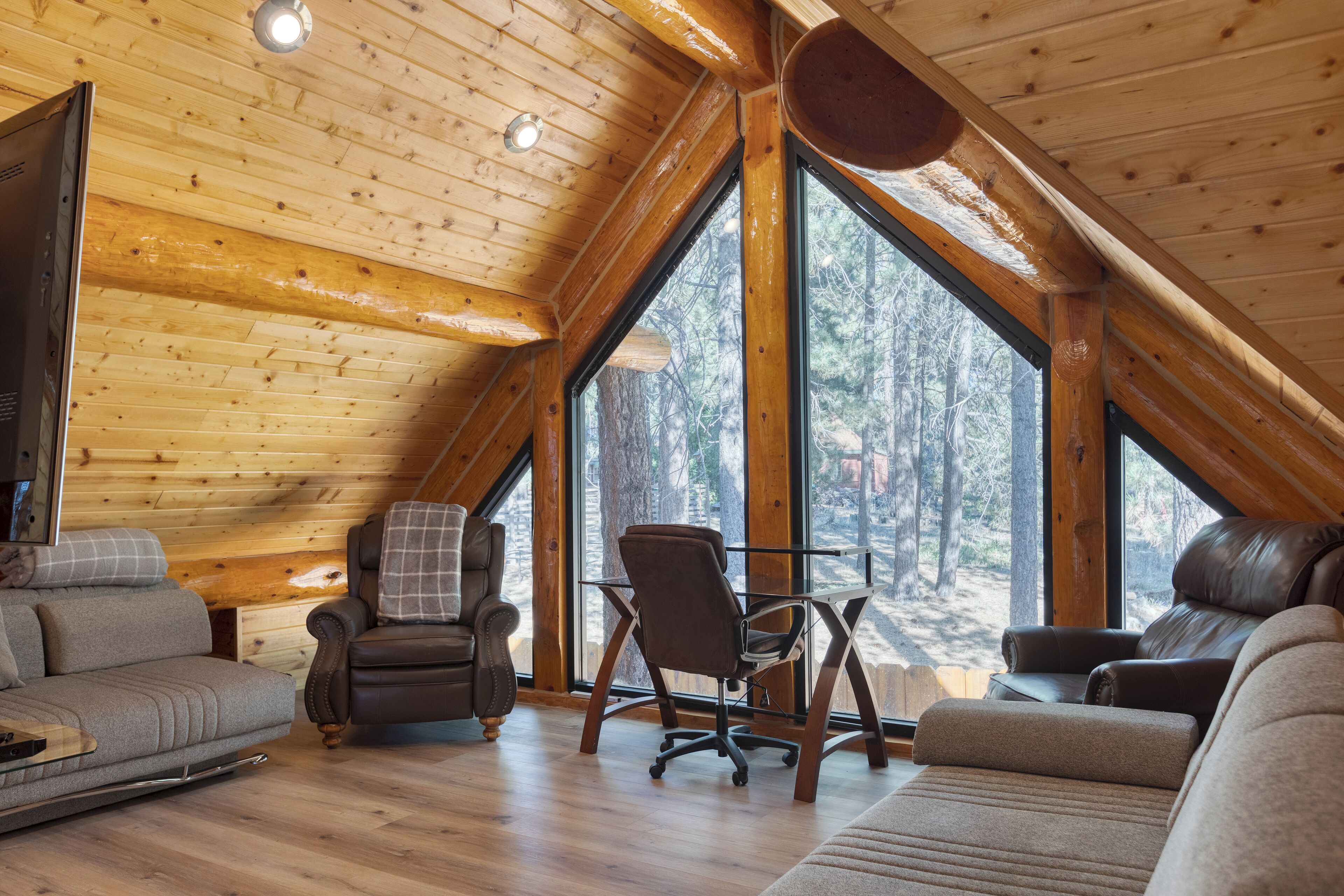 Interior de una cabaña de troncos con un altillo para dormir, suelos de madera y ventanas grandes con vistas al bosque.
