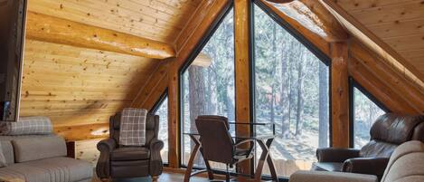 Interior de una cabaña de troncos con un altillo para dormir, suelos de madera y ventanas grandes con vistas al bosque.